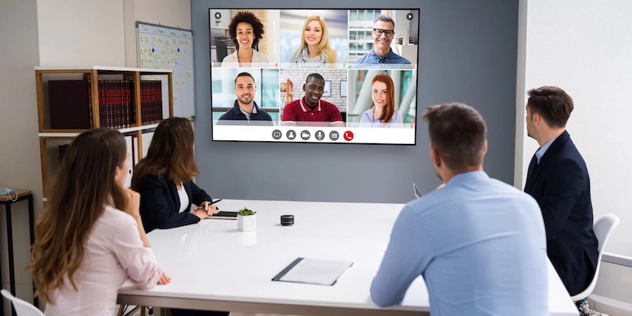 four people sitting around a meeting table, looking at a large TV display with six virtual attendees