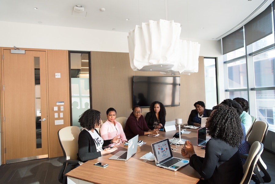 eight women of color sitting around a conference table with laptops and a wall-mounted TV in the background